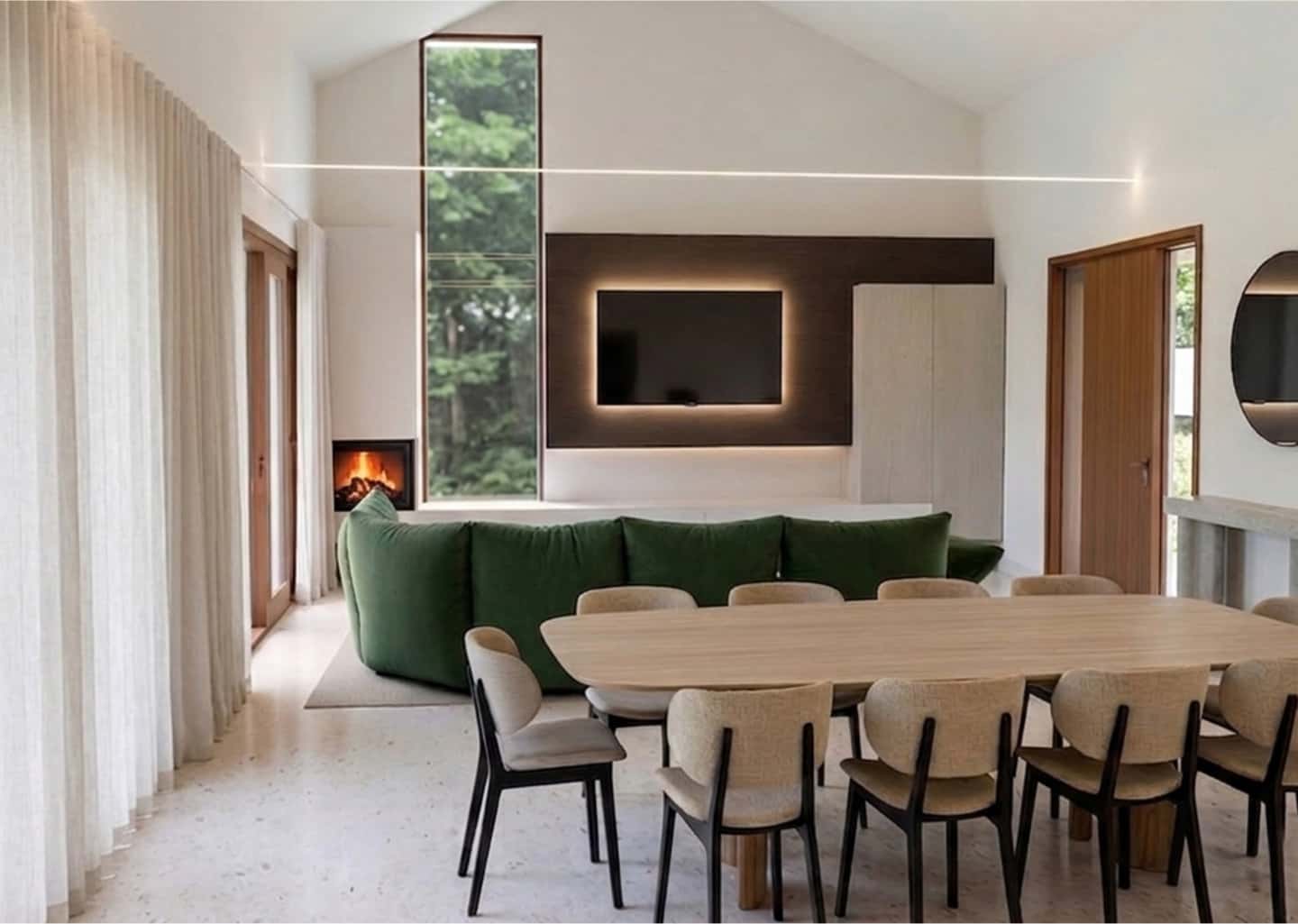 Modern living room with a large green sofa, wooden dining table, chairs, wall-mounted TV, fireplace, tall window, and neutral decor.