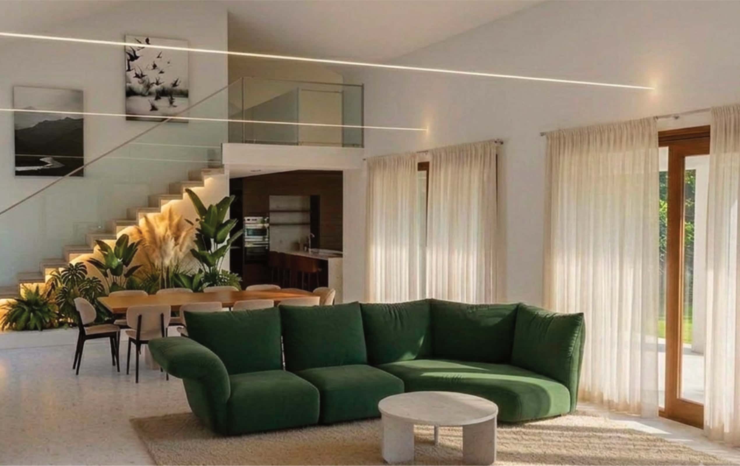 Spacious modern living room with a green sofa, large windows, sheer curtains, indoor plants, a dining table, and staircase leading to a second floor.