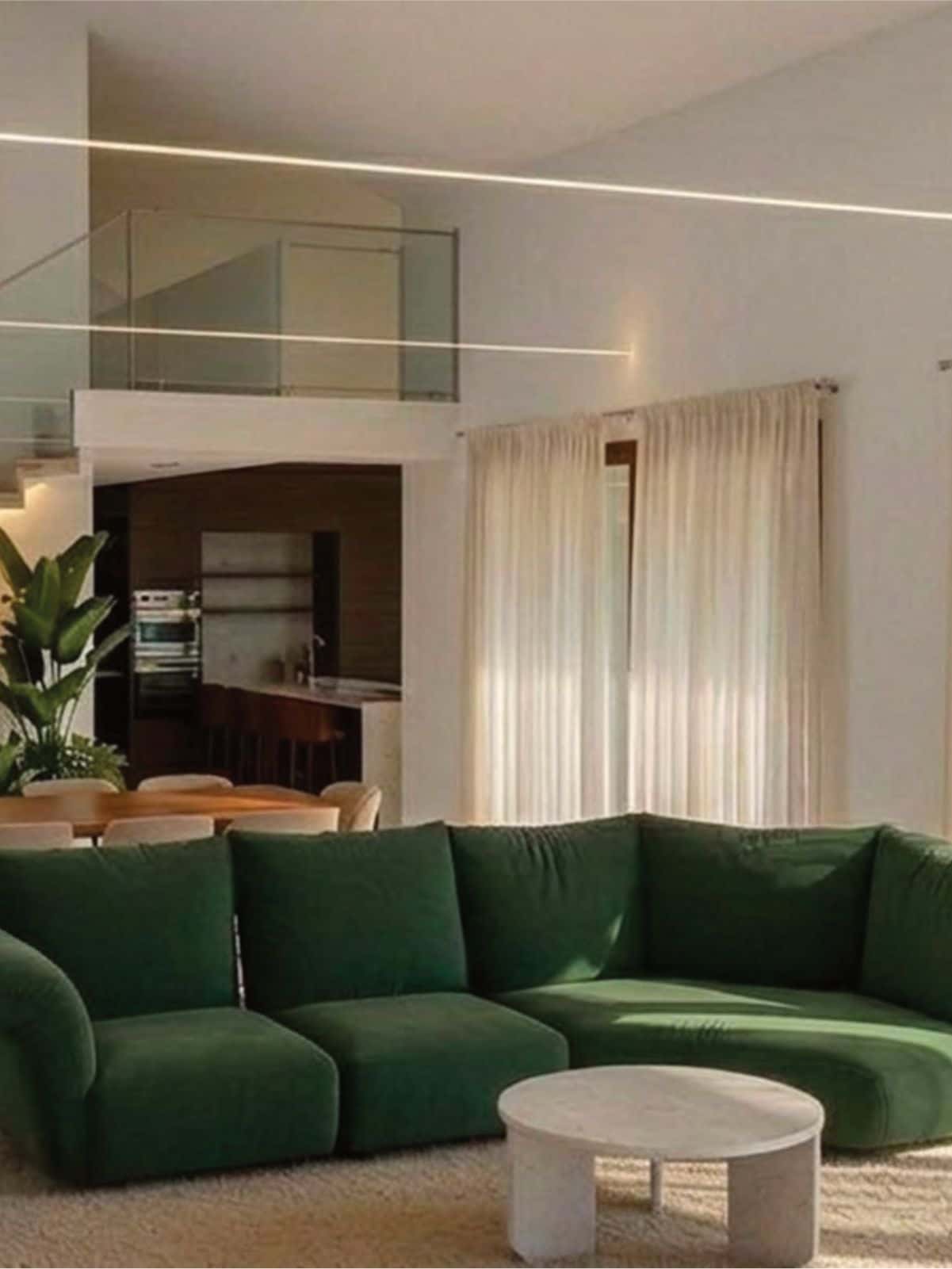 Spacious modern living room with a green sofa, large windows, sheer curtains, indoor plants, a dining table, and staircase leading to a second floor.